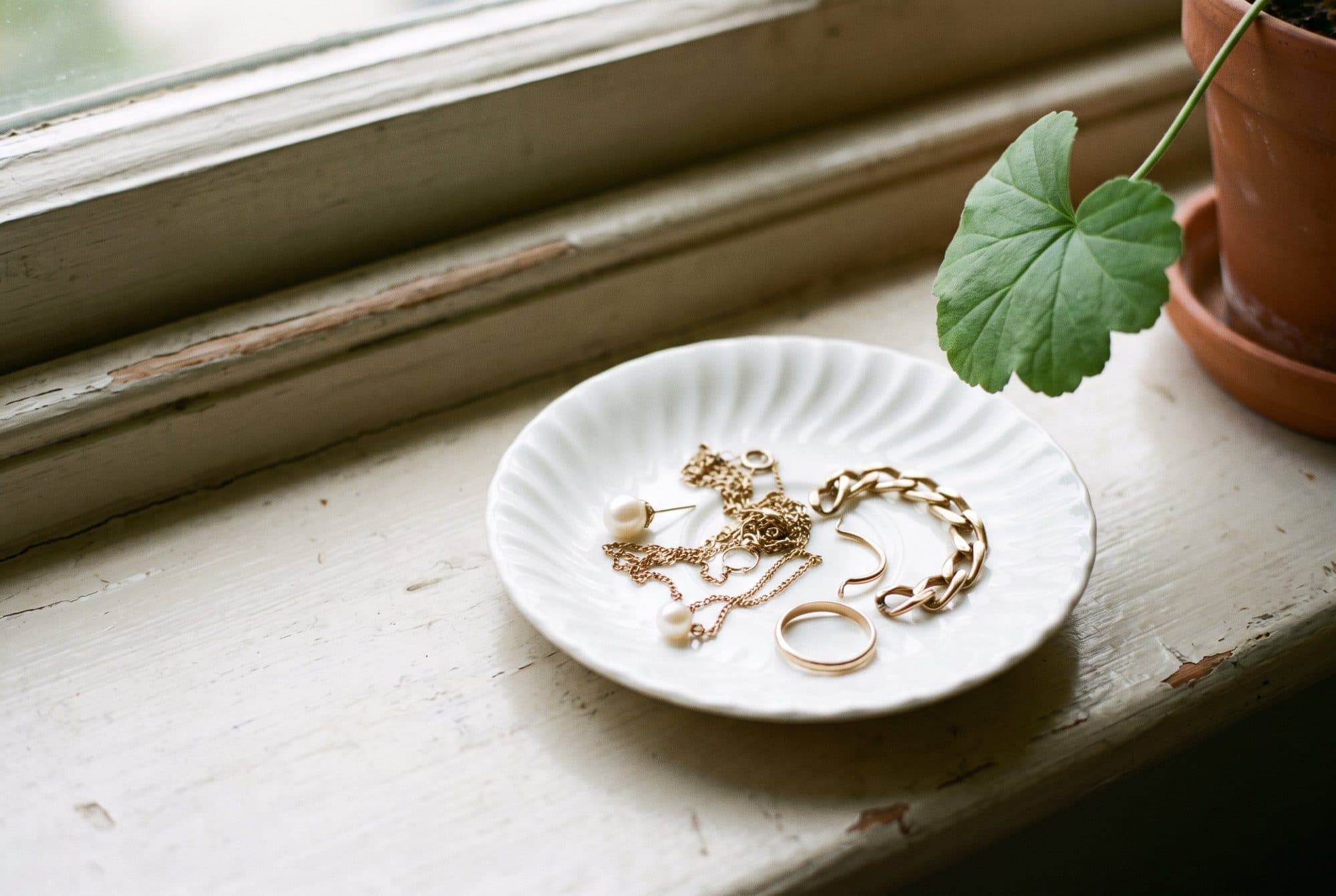 A fluted white saucer on a painted windowsill holding tangled gold chains, odd earrings and two rings.