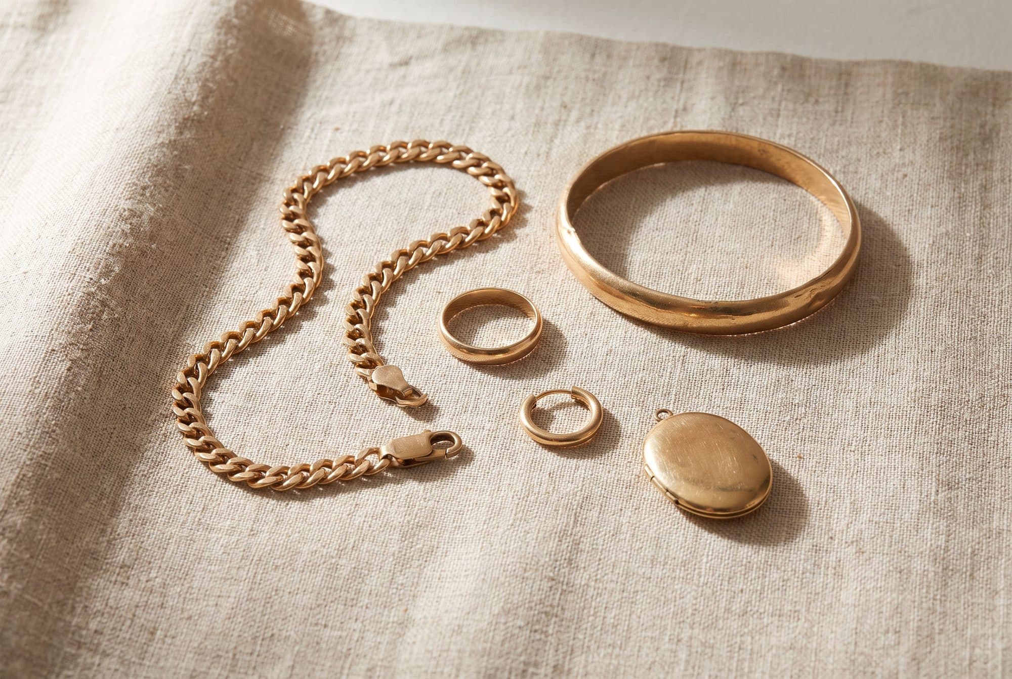 A gold curb chain, a bangle, three rings and an oval locket laid out on plain linen.