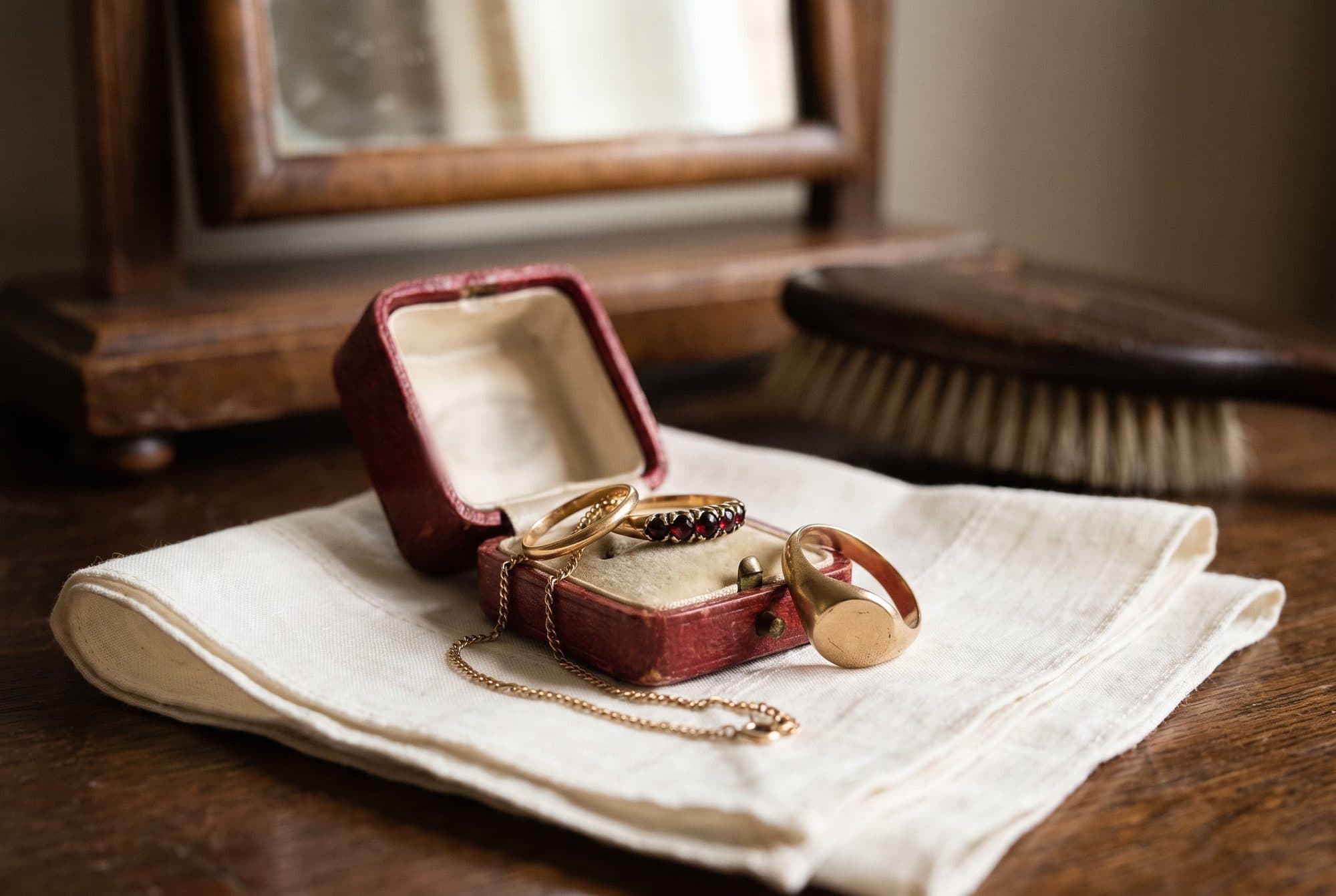 A worn red leather ring box on a folded linen cloth in front of a dressing-table mirror, holding rings, with a signet ring and a fine chain beside it.