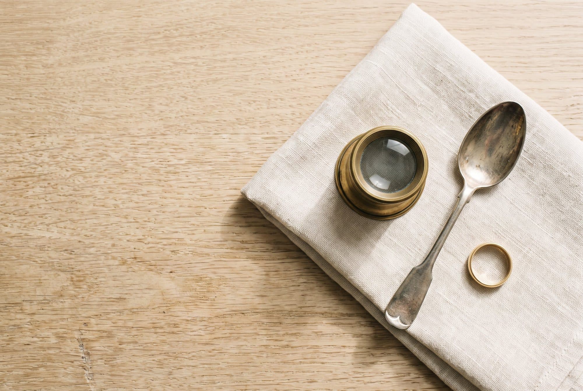 A brass-rimmed loupe, a silver teaspoon and a gold ring on a folded linen cloth on a pale oak table.