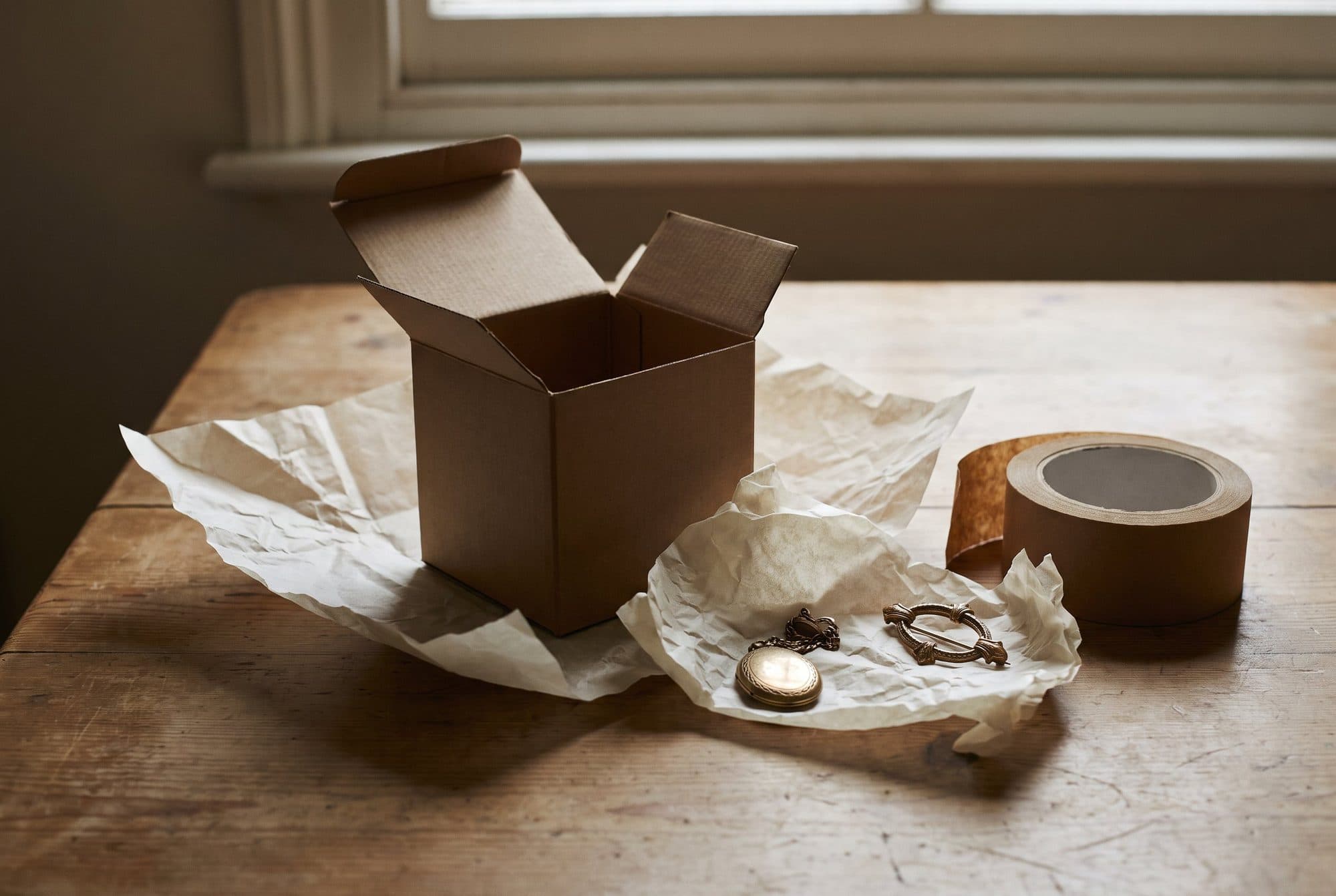 A small plain cardboard box being packed on a table, with tissue paper, a roll of parcel tape and a few pieces of jewellery.