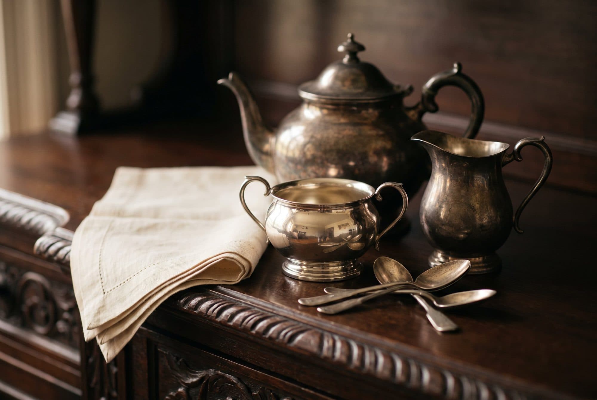 A silver teapot, sugar bowl and cream jug with a few teaspoons and a folded napkin on a carved wooden sideboard.
