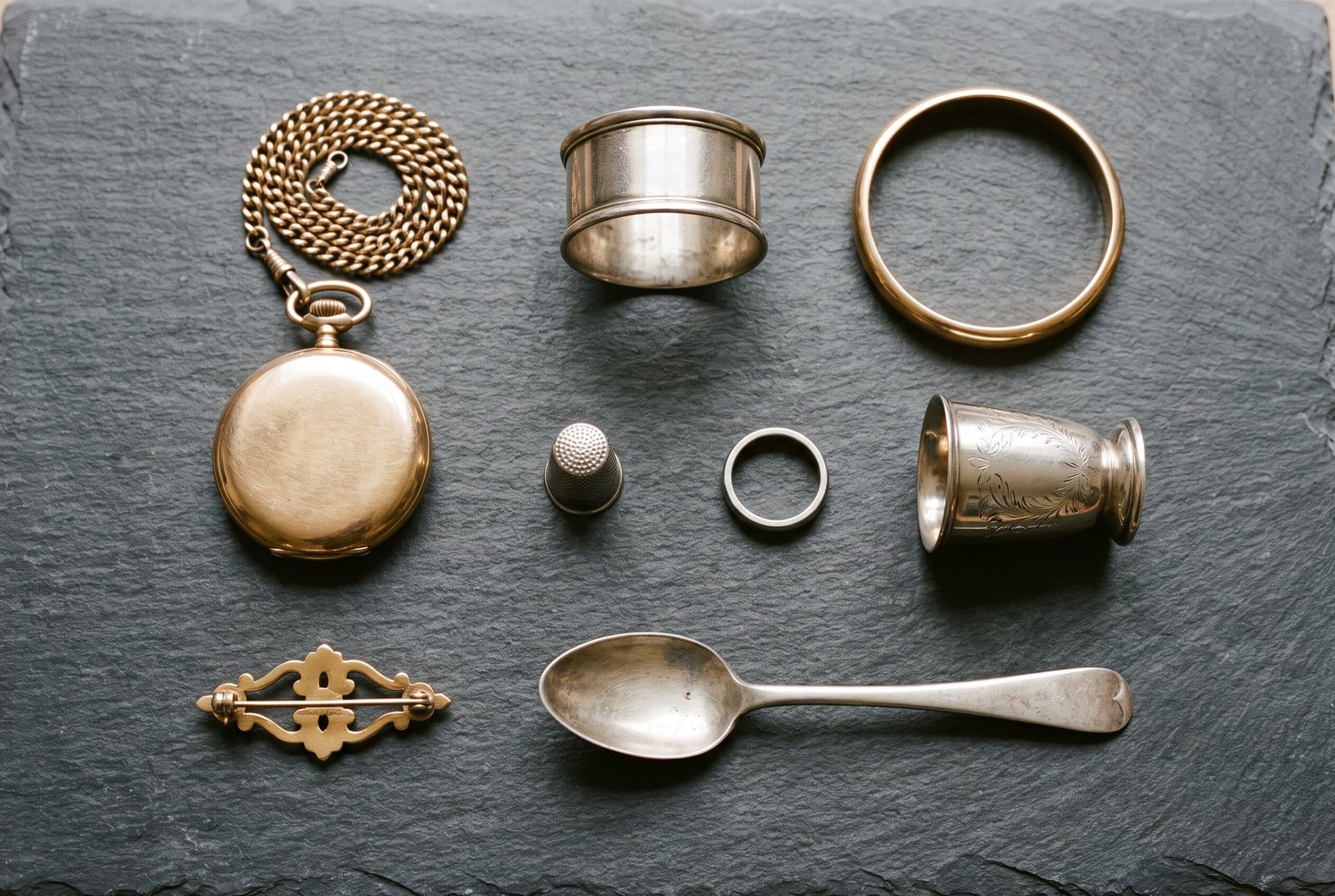 A pocket watch on its chain, a bangle, a napkin ring, a thimble, a ring, a small beaker, a brooch and a spoon laid out on a slate.